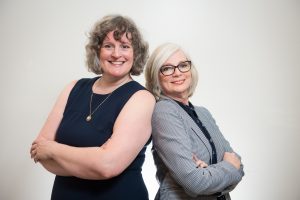 Angela Crocker wearing a sleeveless navy blue dress standing back to back with Vicki McLeod wearing a navy blouse and checkered blazer.