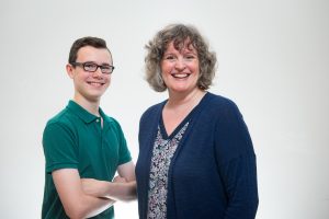 Sean Crocker and his mother, author Angela Crocker. Sean stands with his arms folded across his chest is wearing a green polo shirt. Angela smiling next to him is wearing a floral top and navy blue cardigan.