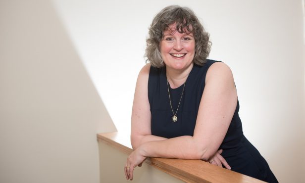 Angela Crocker, author and educator, wearing a navy blue sleeveless dress leans casually on a low wall smiling at the camera.