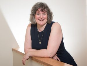 Angela Crocker, author and educator, wearing a navy blue sleeveless dress leans casually on a low wall smiling at the camera.