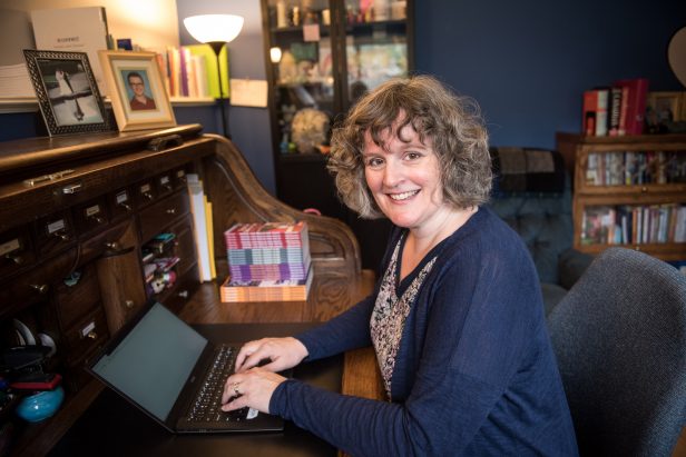Author Angela Crocker seated at her rolltop desk typing on her lapt top. Books in the background.