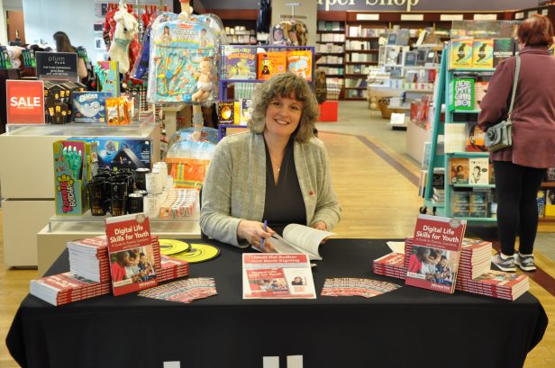 Author Angela Crocker seated at a book signing table in a bookstore. Copies of Digital Life Skills for Youth are on the table.