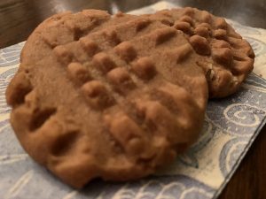 Two peanut butter cookies sit on a blue and white paper napin. The cookies have a cross hatch pattern made with the tines of a fork.